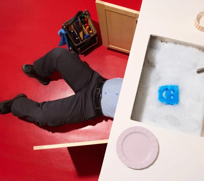 Person fixing plumbing under a kitchen sink, partially visible with legs sticking out, with a tool bag nearby and a smiley face sponge floating in a soapy sink.