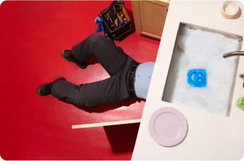 Person fixing plumbing under a kitchen sink, partially visible with legs sticking out, with a tool bag nearby and a smiley face sponge floating in a soapy sink.